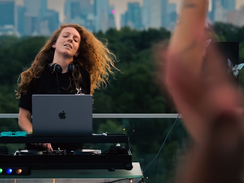 Jess DJing on a roof deck with treetops and a skyline in the background.