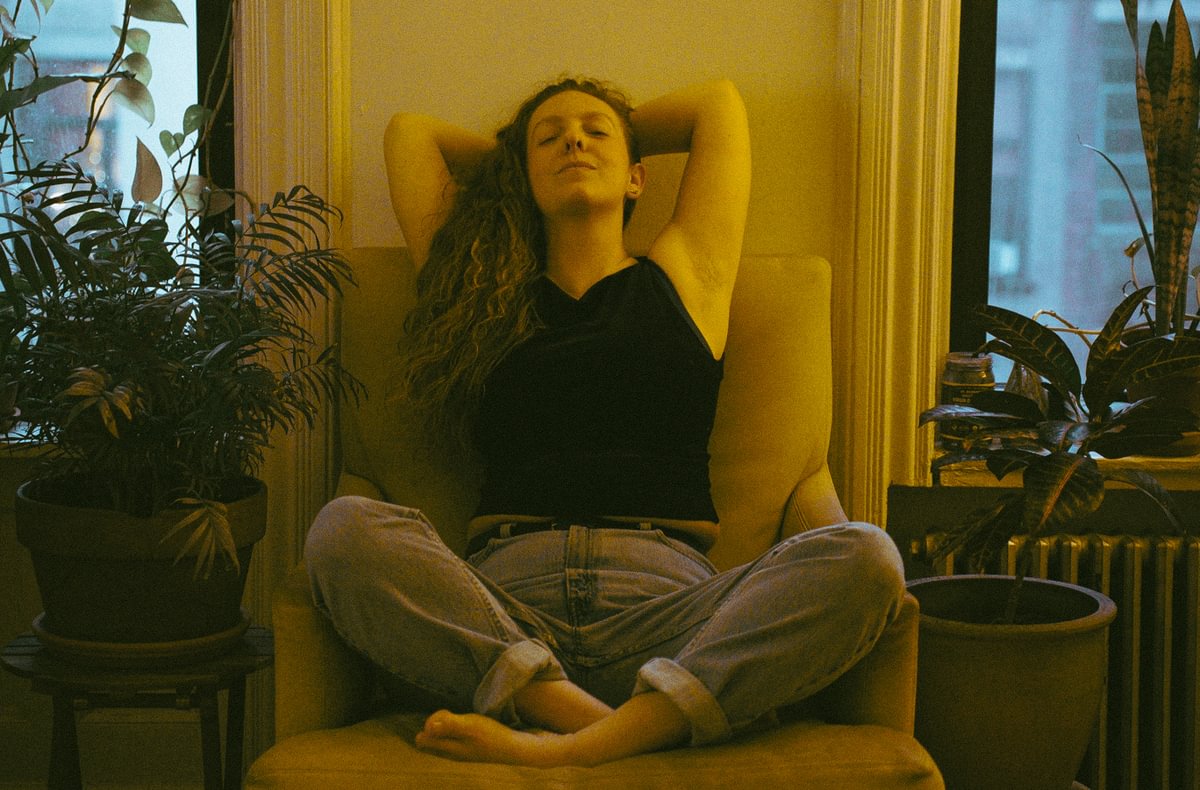 Jess Here sitting lotus style on a plush chair, arms crossed behind her head, and relaxing with her eyes closed.